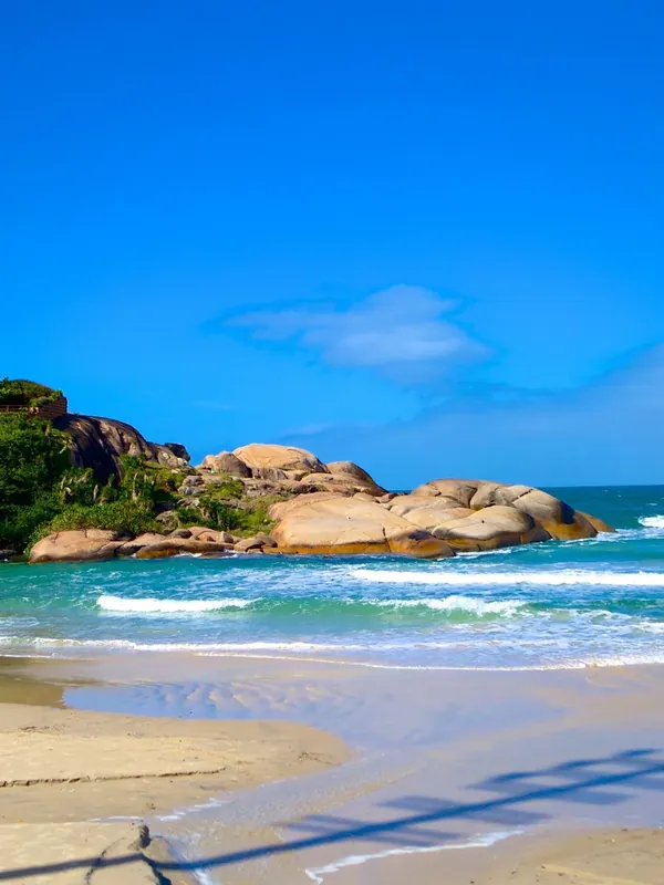 Praia da Joaquina, com areia clara, ondas e o costão de pedras sob céu azul.