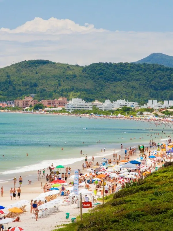 Praia de Jurerê cheia de banhistas e guarda-sóis, com mar calmo e morros verdes ao fundo.